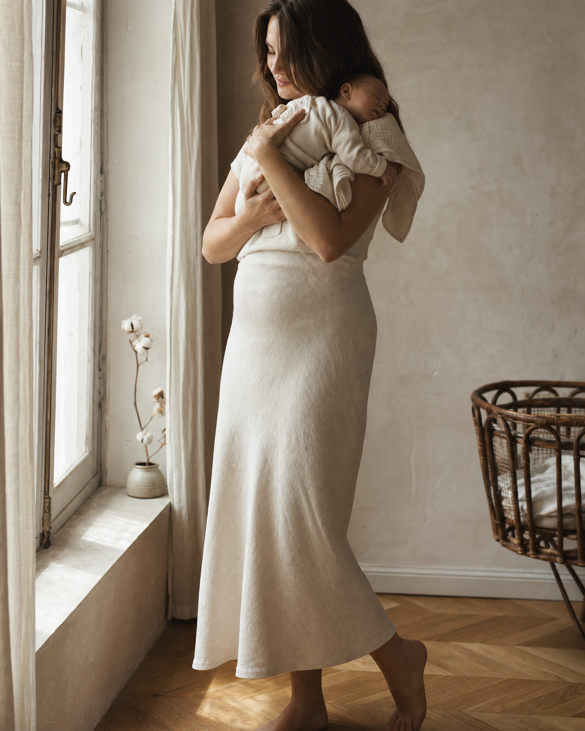 Jeune maman en robe de lin tenant son nouveau-né endormi près d’une fenêtre